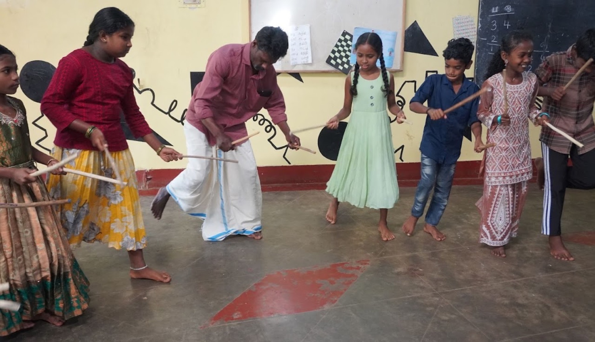 Adivasi children and a teacher doing a stick-dance (kolattam) inside a classroom