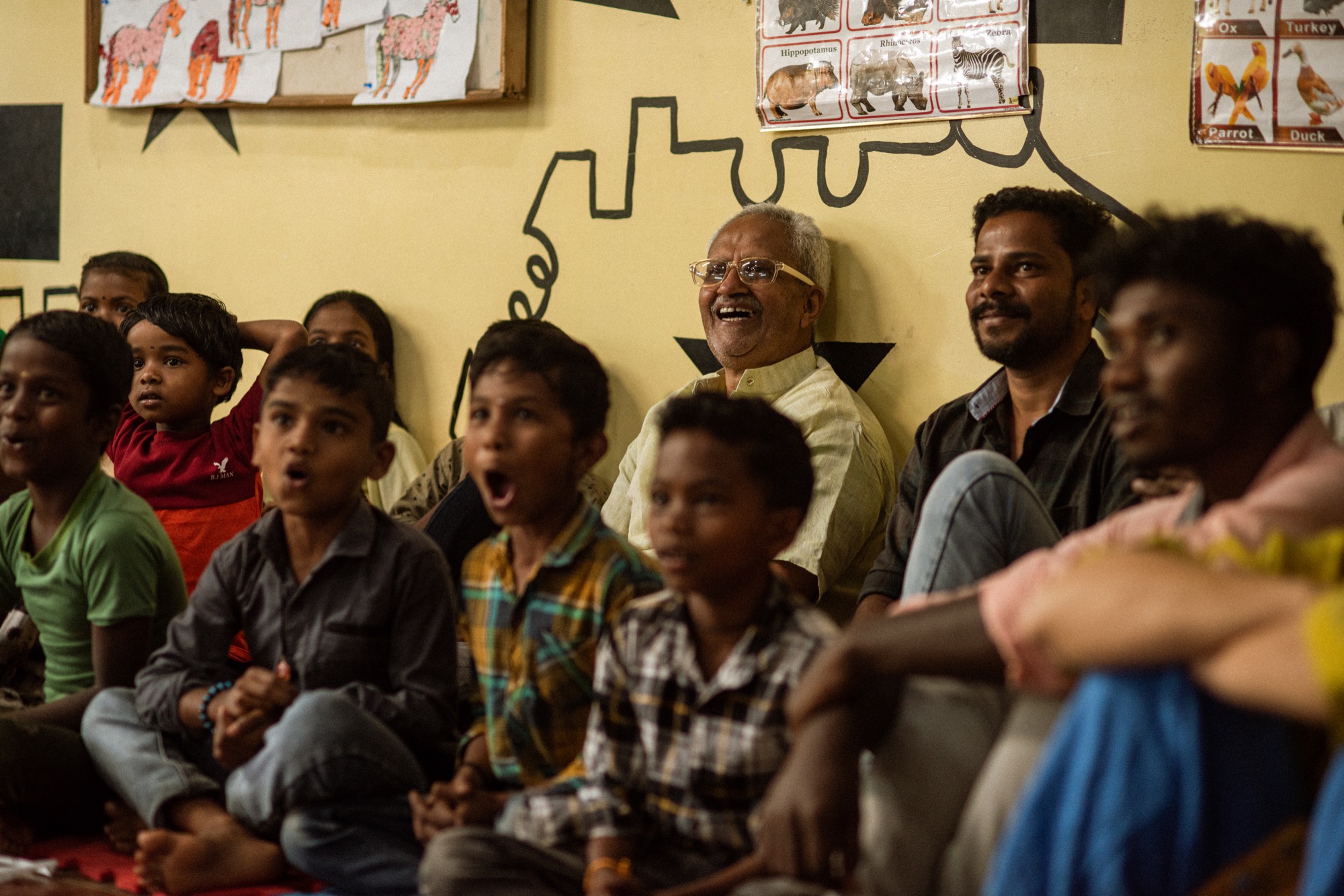 Adivasi children and elders seated together watching a performance inside a classroom, an elder man laughing