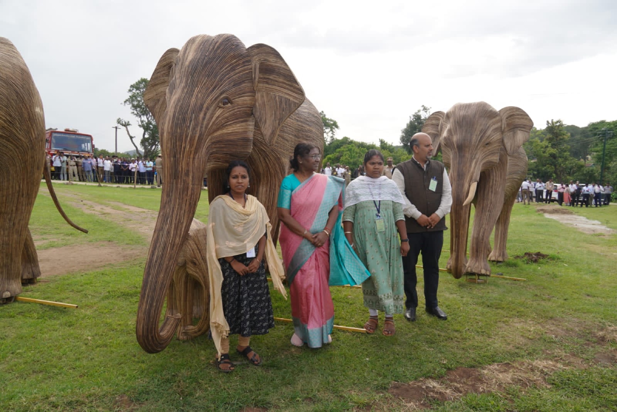 Adivasi community members standing with President Droupadi Murmu beside life-size bamboo elephant sculptures on a green field