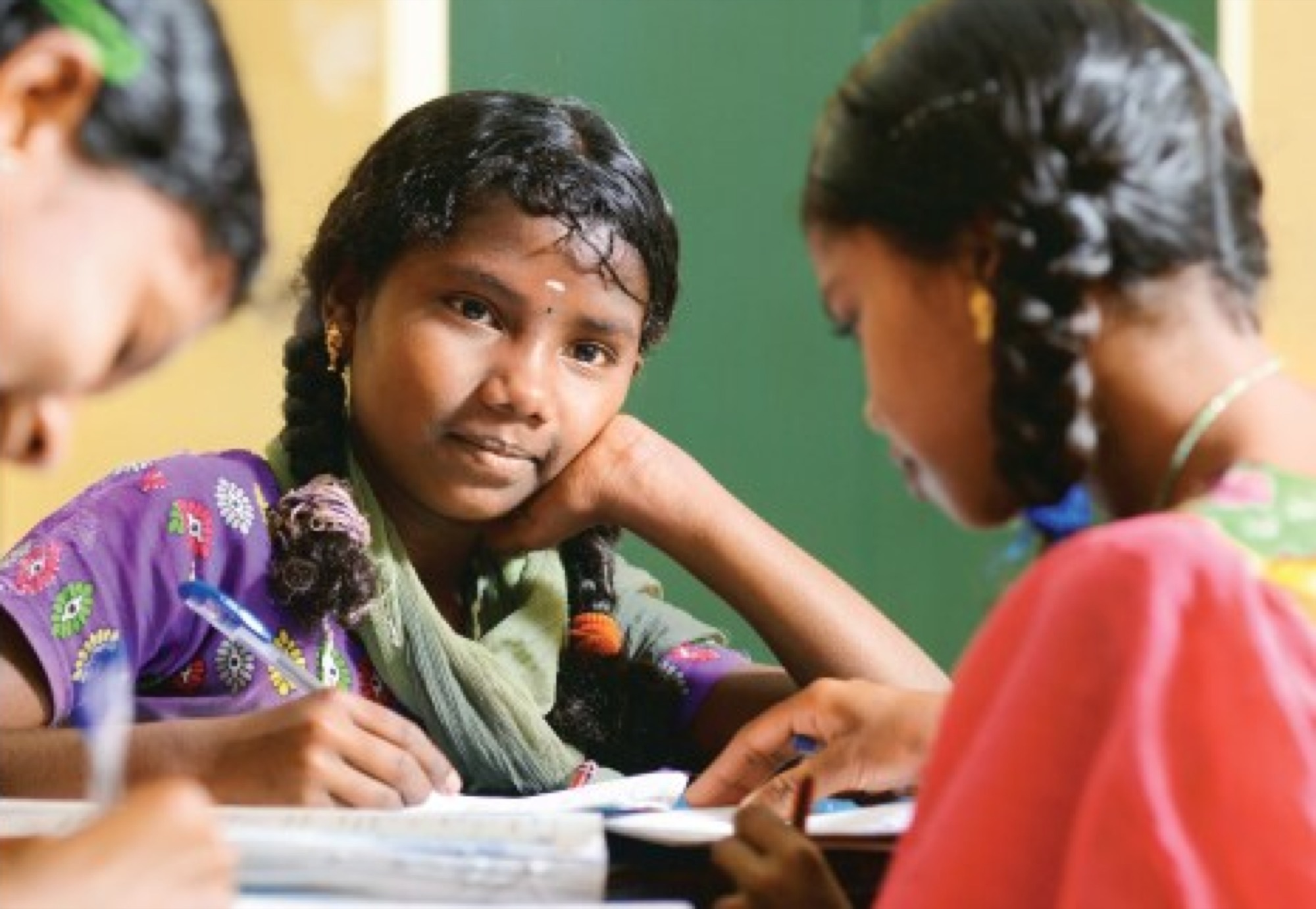 Adivasi girl in a classroom looking up thoughtfully while writing with two other students