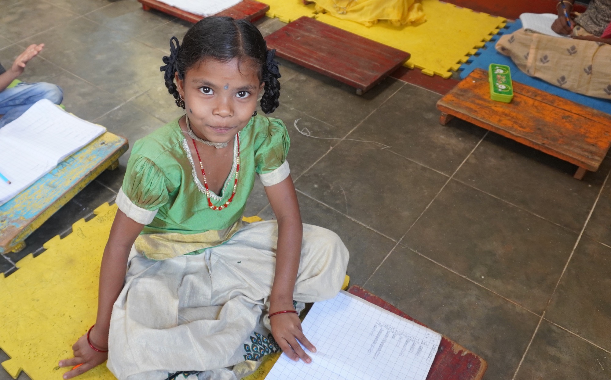 Adivasi girl in a green silk dress seated on the floor with her notebook, looking up at camera