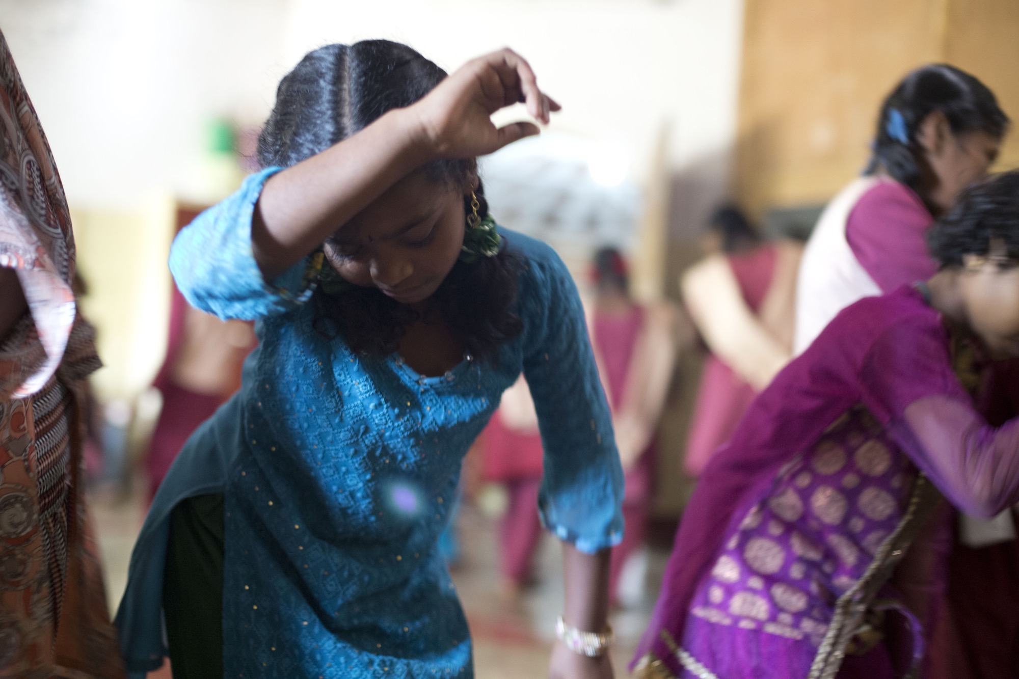 Adivasi girls and young women dancing together indoors, a girl in a blue kurta mid-movement in the foreground