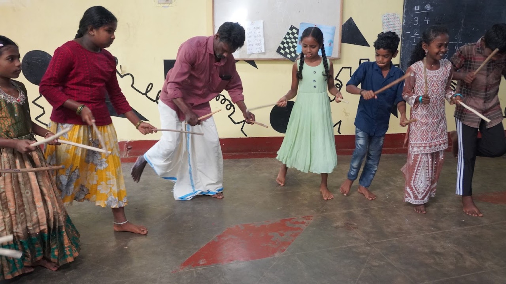Children and a man performing the kolattam stick-dance with wooden sticks in a learning-centre hall