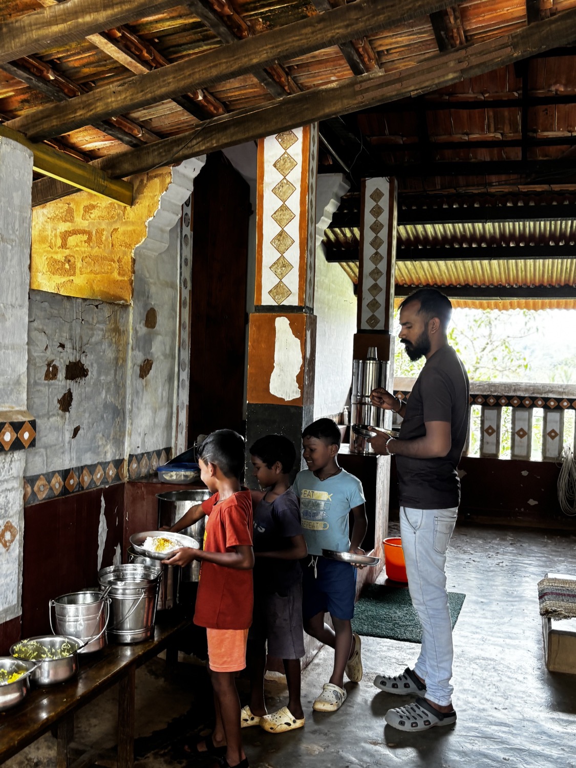 Children lining up at a serving counter for a meal while a man serves, on a decorated mud-wall veranda