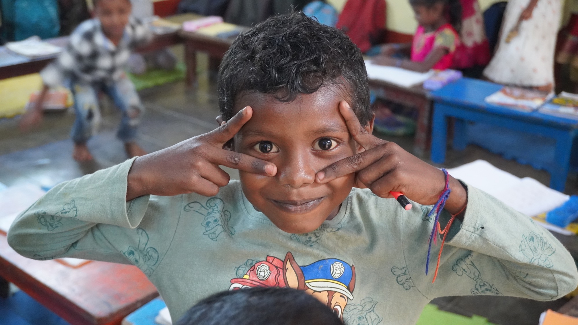Close-up portrait of a playful Adivasi boy framing his eyes with his fingers in a classroom
