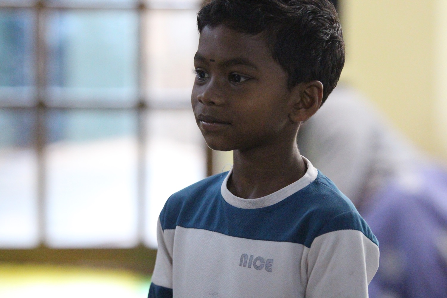 Crisp close portrait of an Adivasi boy in a blue-and-white shirt, soft window light