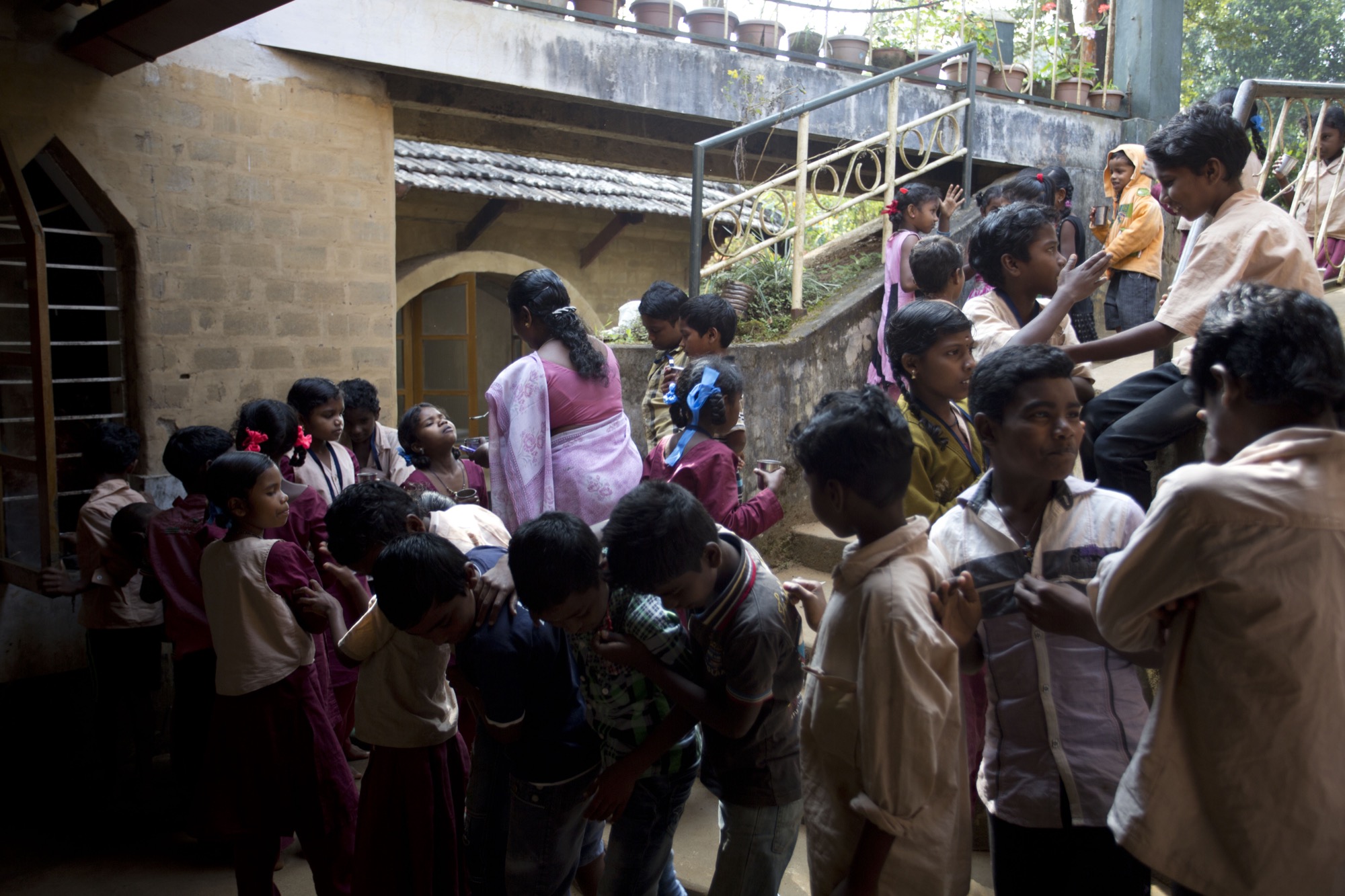 Crowd of Adivasi children and a teacher in a pink sari moving through the open courtyard outside the school building