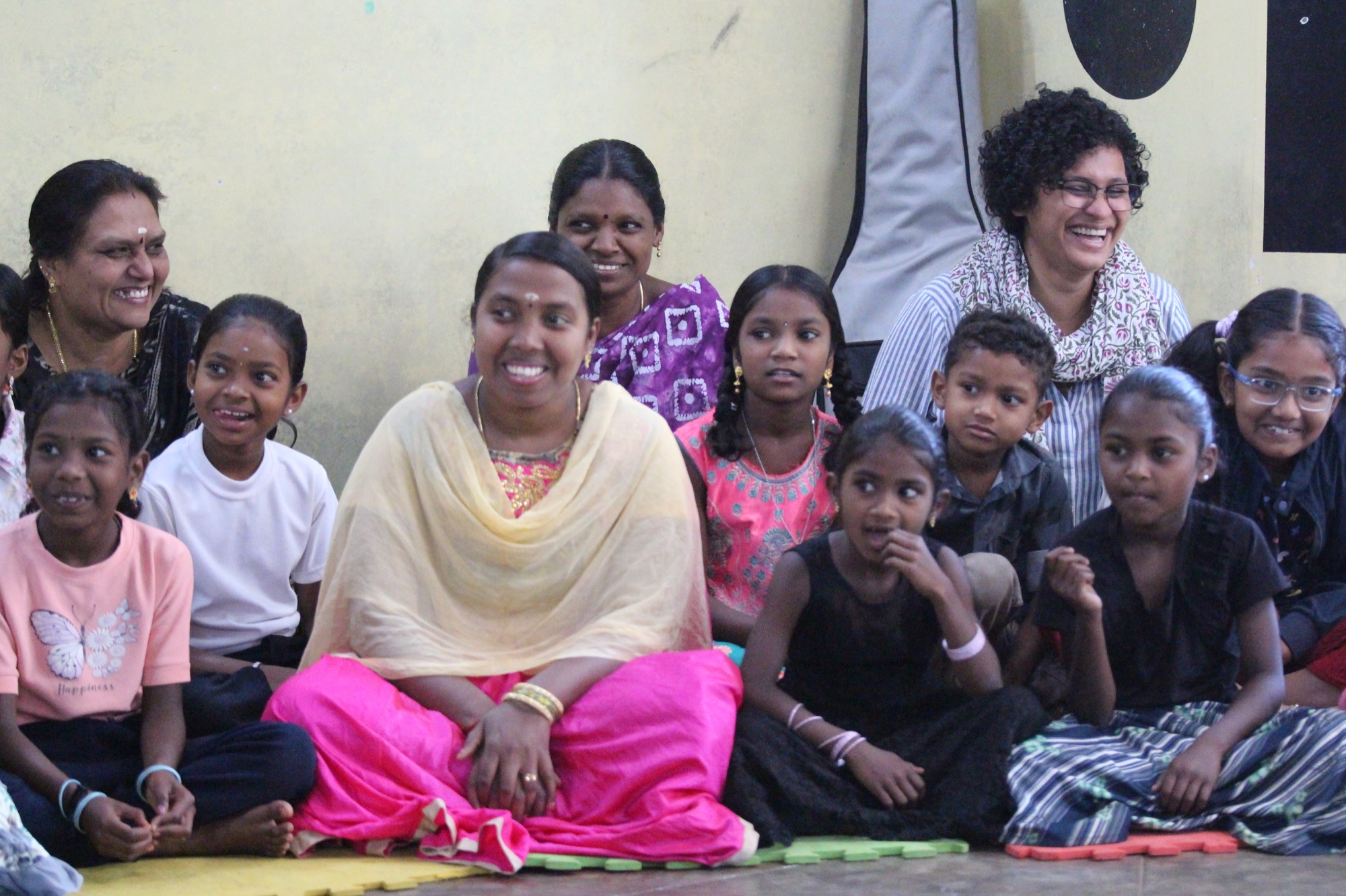 A group of Adivasi women teachers and mothers seated with girls, all smiling warmly