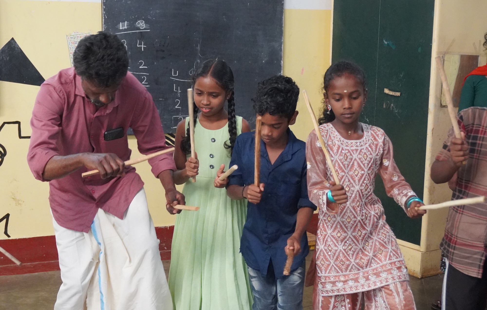 Male teacher in maroon shirt and dhoti leading children in a stick-dance lesson in front of a chalkboard