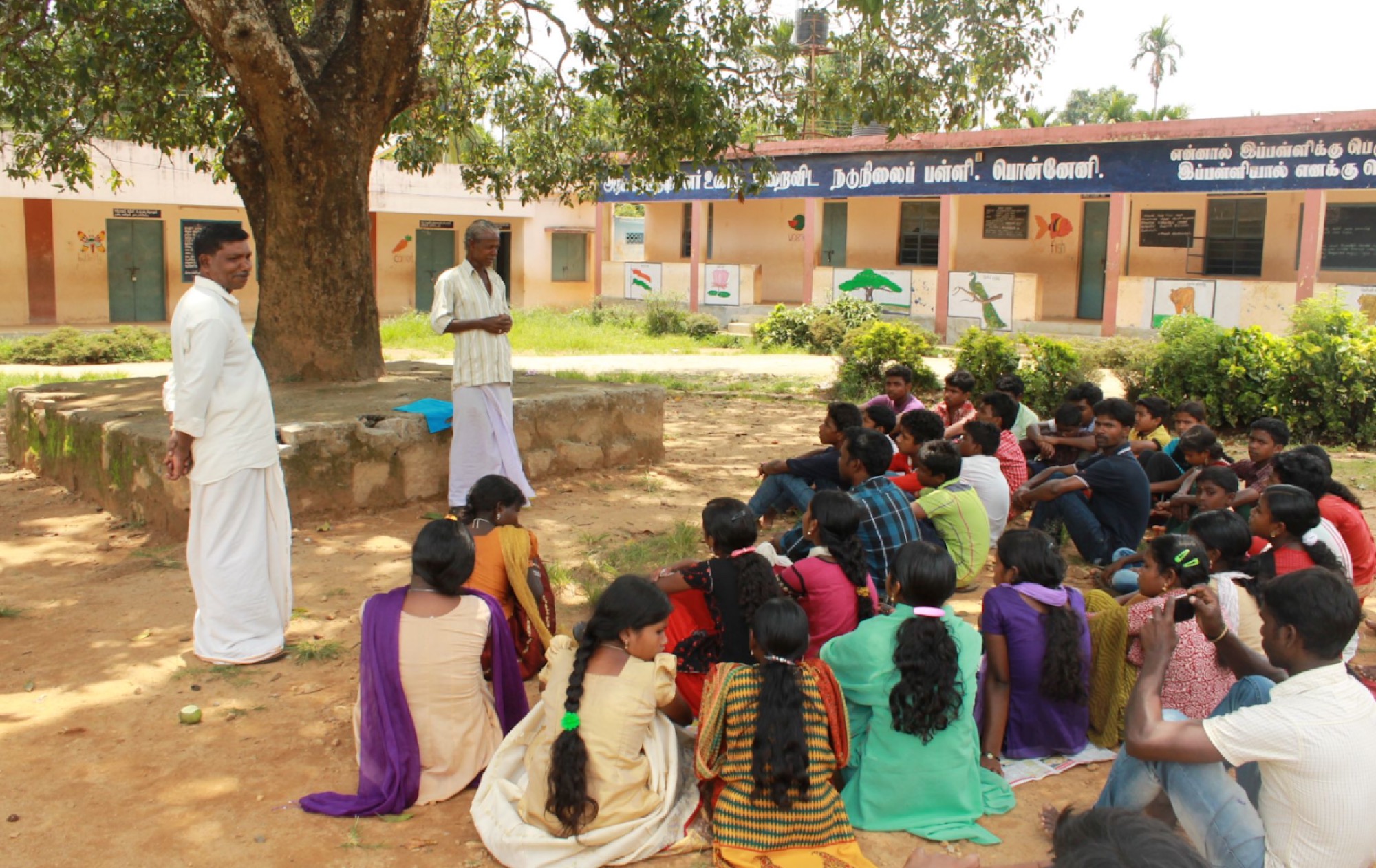 Outdoor camp session, a man addressing a large group of children seated on the ground under a tree at the school