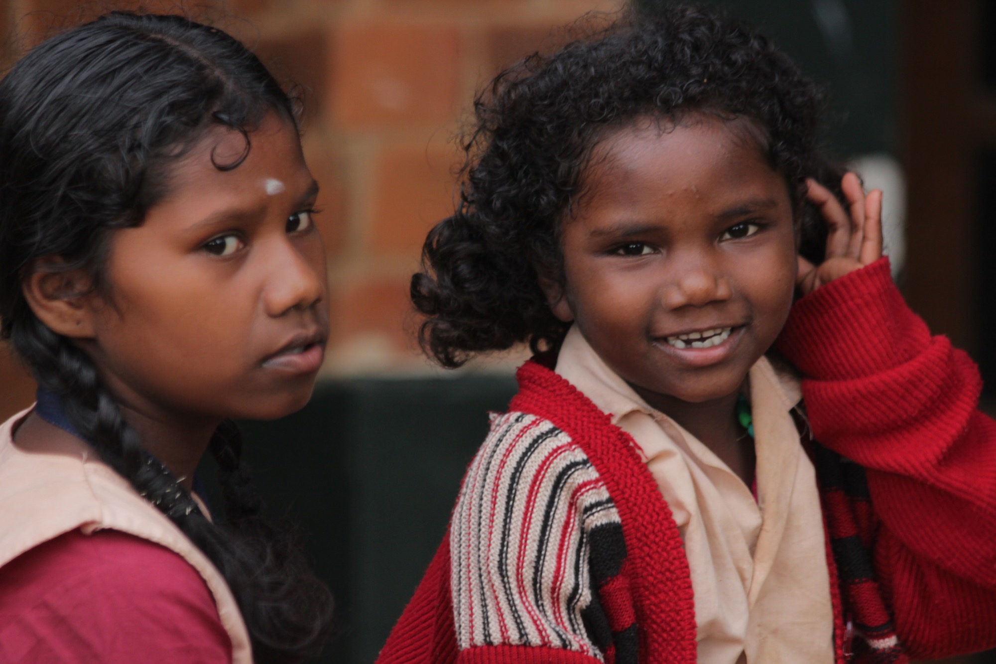 Portrait of two Adivasi children, a curly-haired girl smiling beside an older girl, against a brick wall