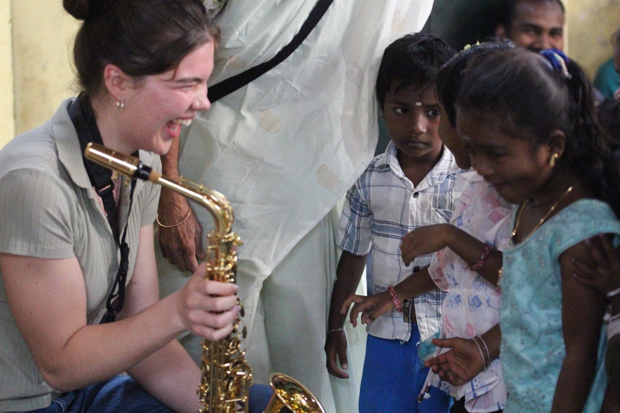 Saxophonist crouching to play for a row of small Adivasi children watching closely
