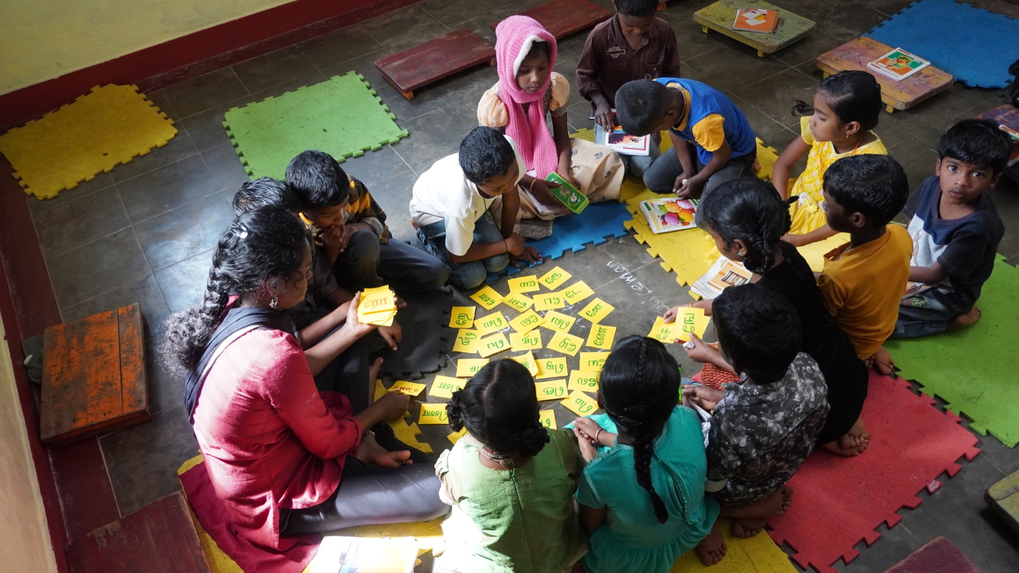Teacher and children sitting in a circle on a classroom floor doing a Tamil word-card learning activity