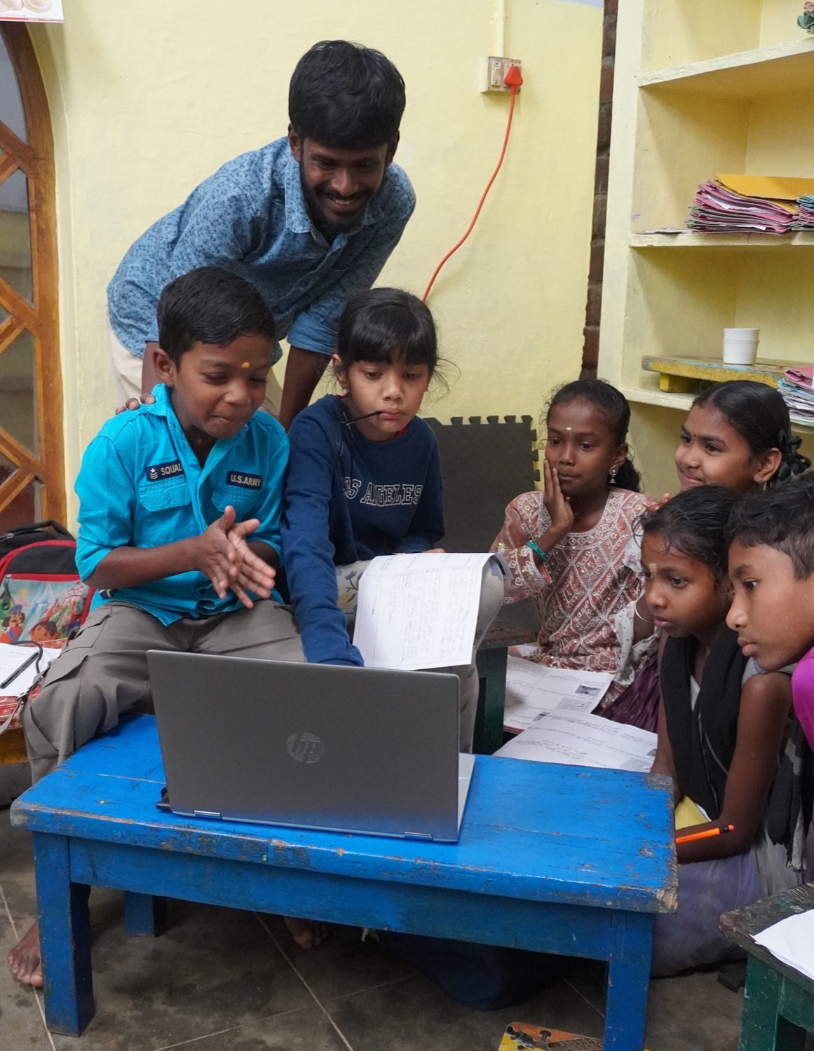 Teacher standing over a blue table guiding Adivasi children using a laptop, kids gathered around attentively