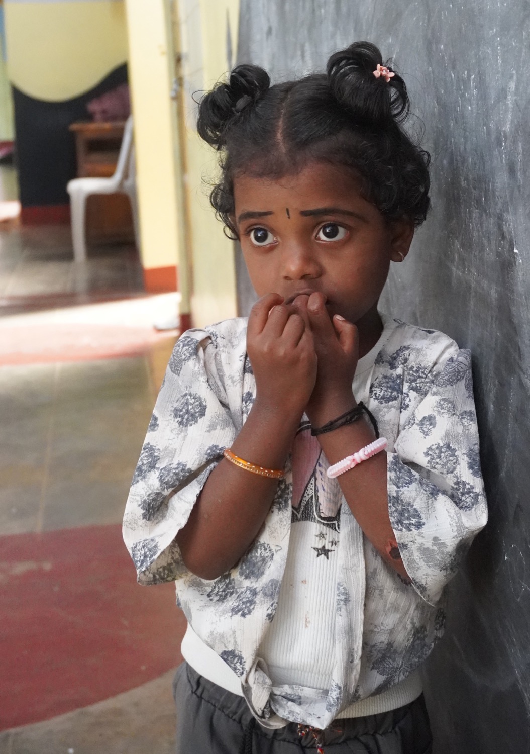 Tender portrait of a small Adivasi girl with twin hair-buns standing against a chalkboard wall, hands to mouth