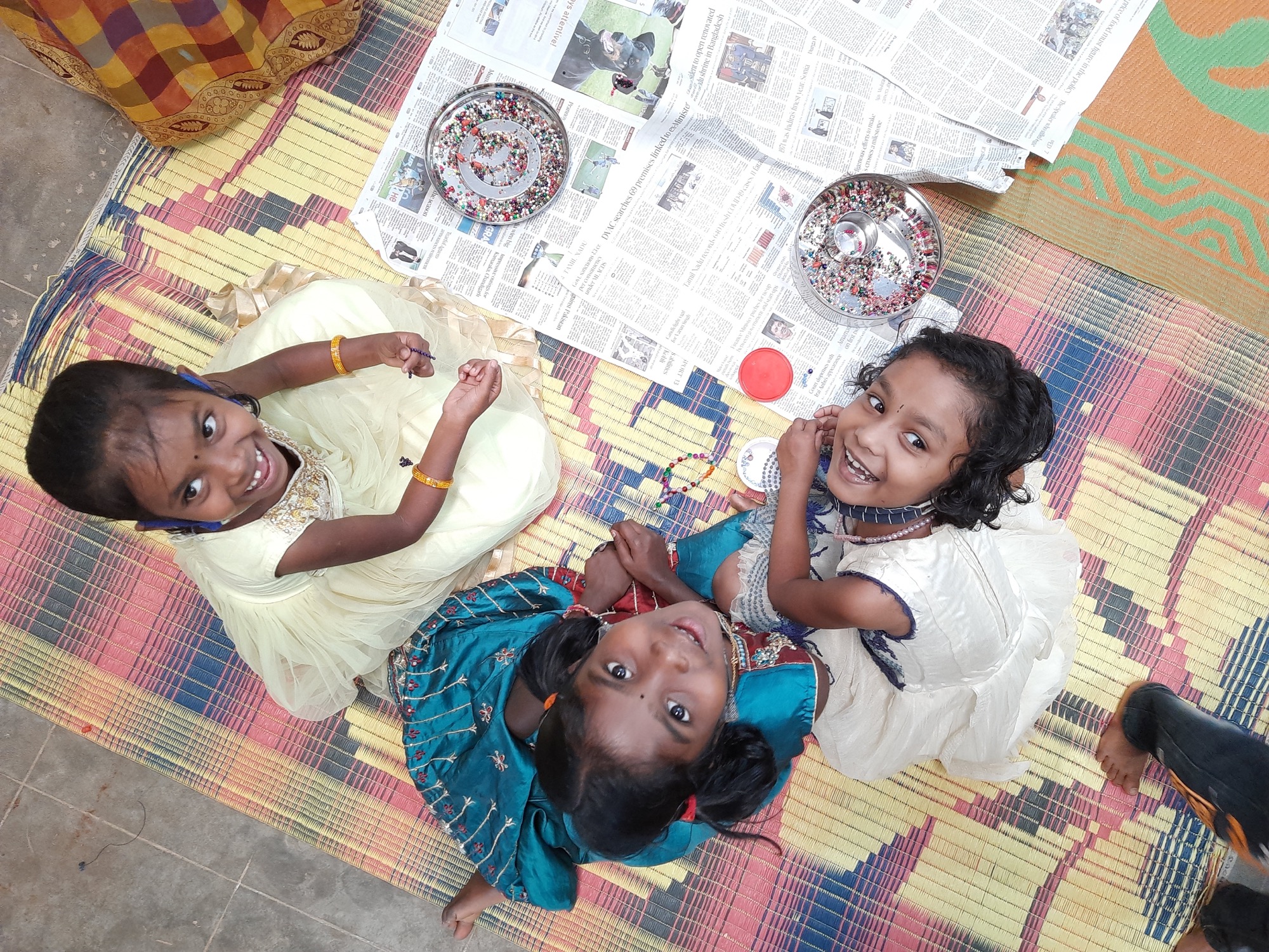 Three young Adivasi girls smiling up at the camera during a craft activity on a mat