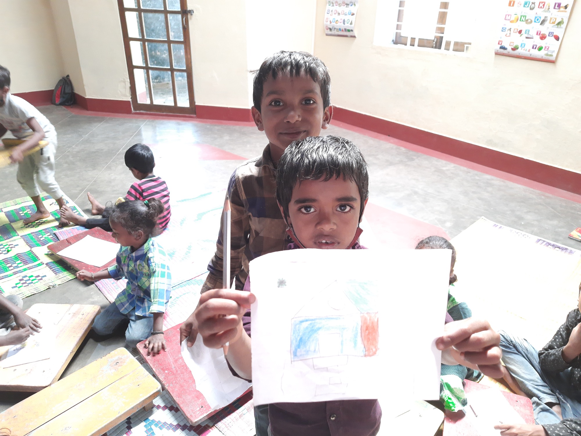 Two boys in a classroom, one proudly holding up his crayon drawing