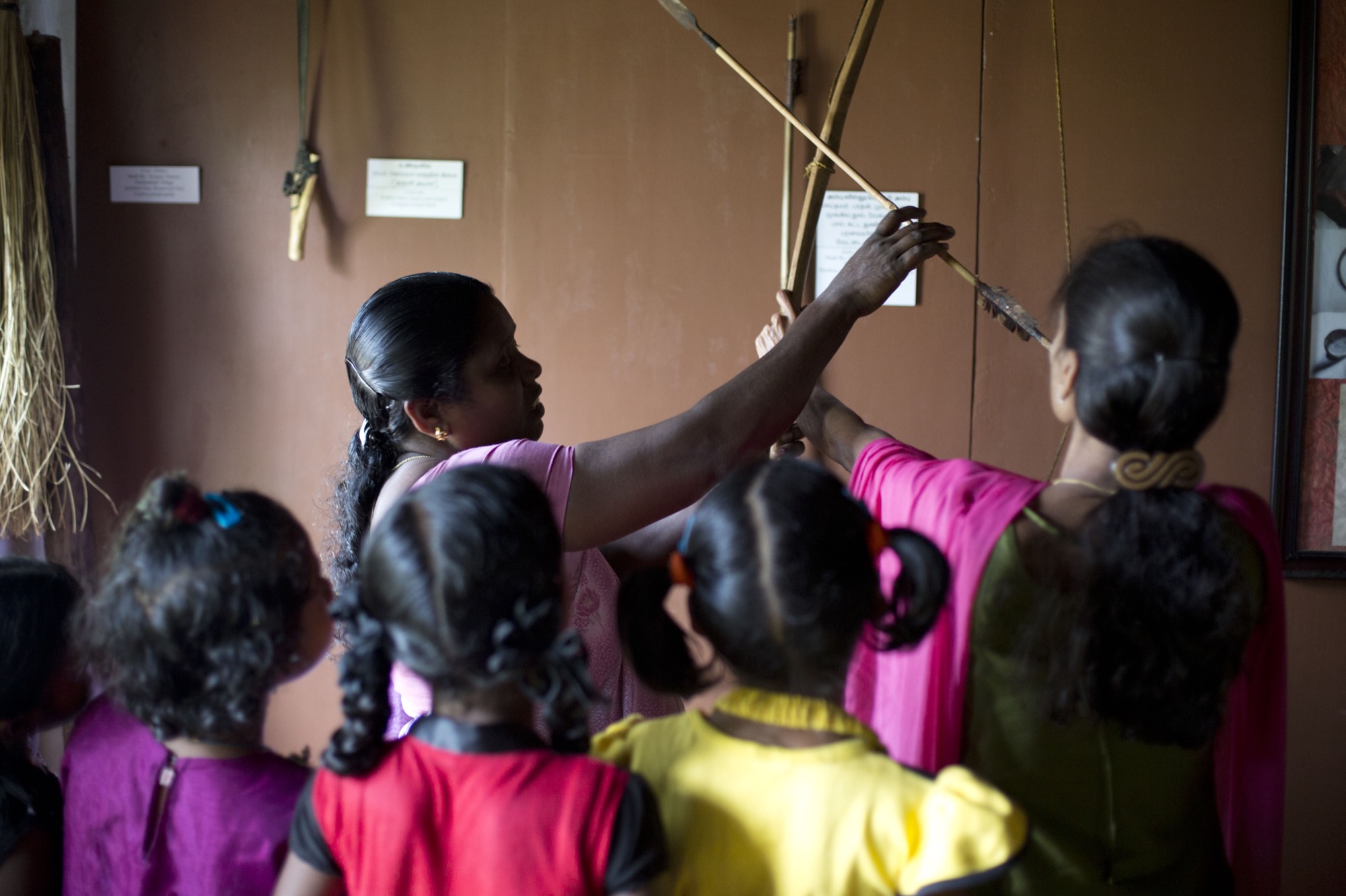 Two women demonstrating bamboo and reed materials on a display wall to a cluster of small girls in bright dresses