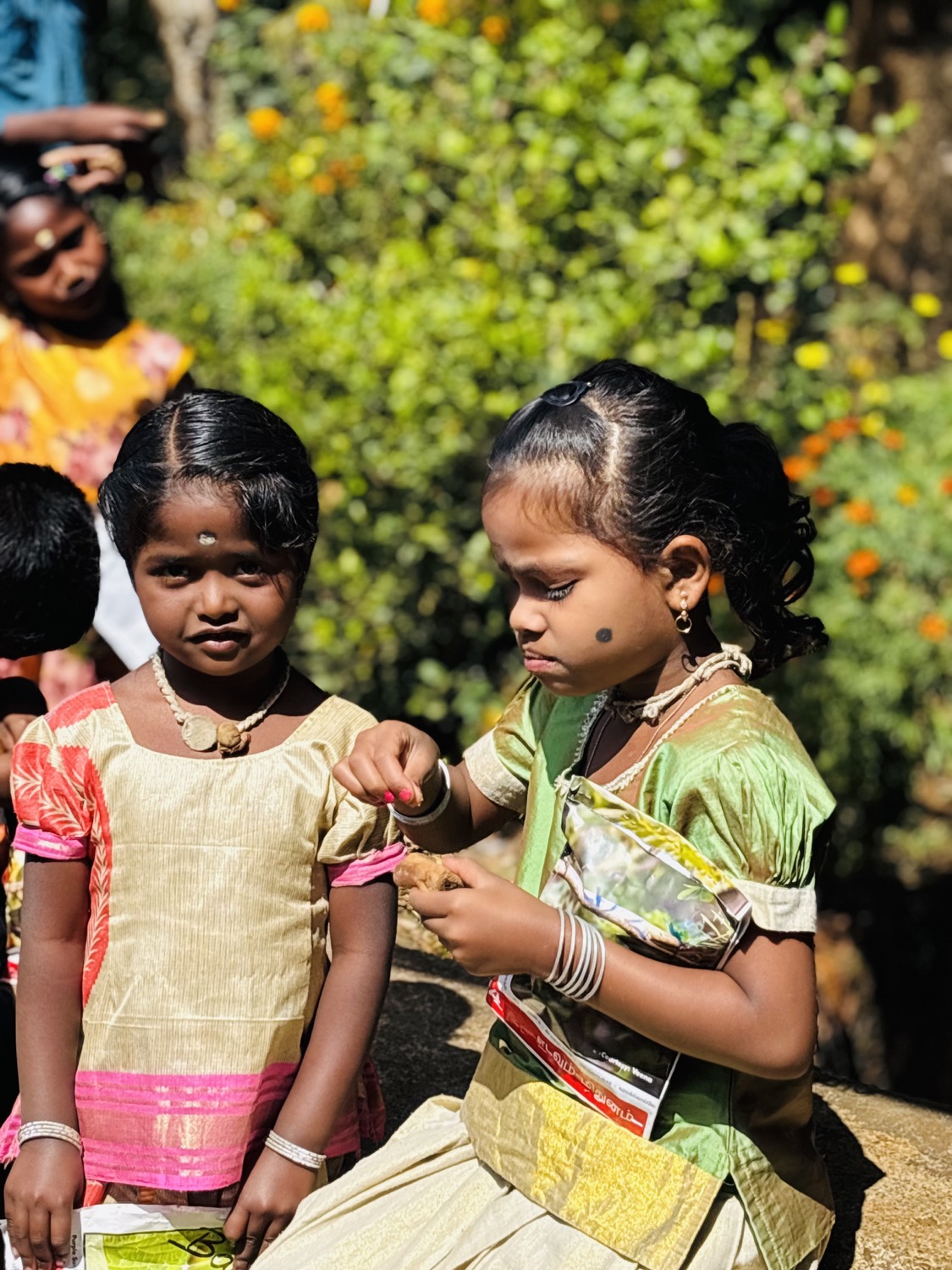 Two young Adivasi girls in traditional gold-and-pink dresses outdoors amid marigold blooms