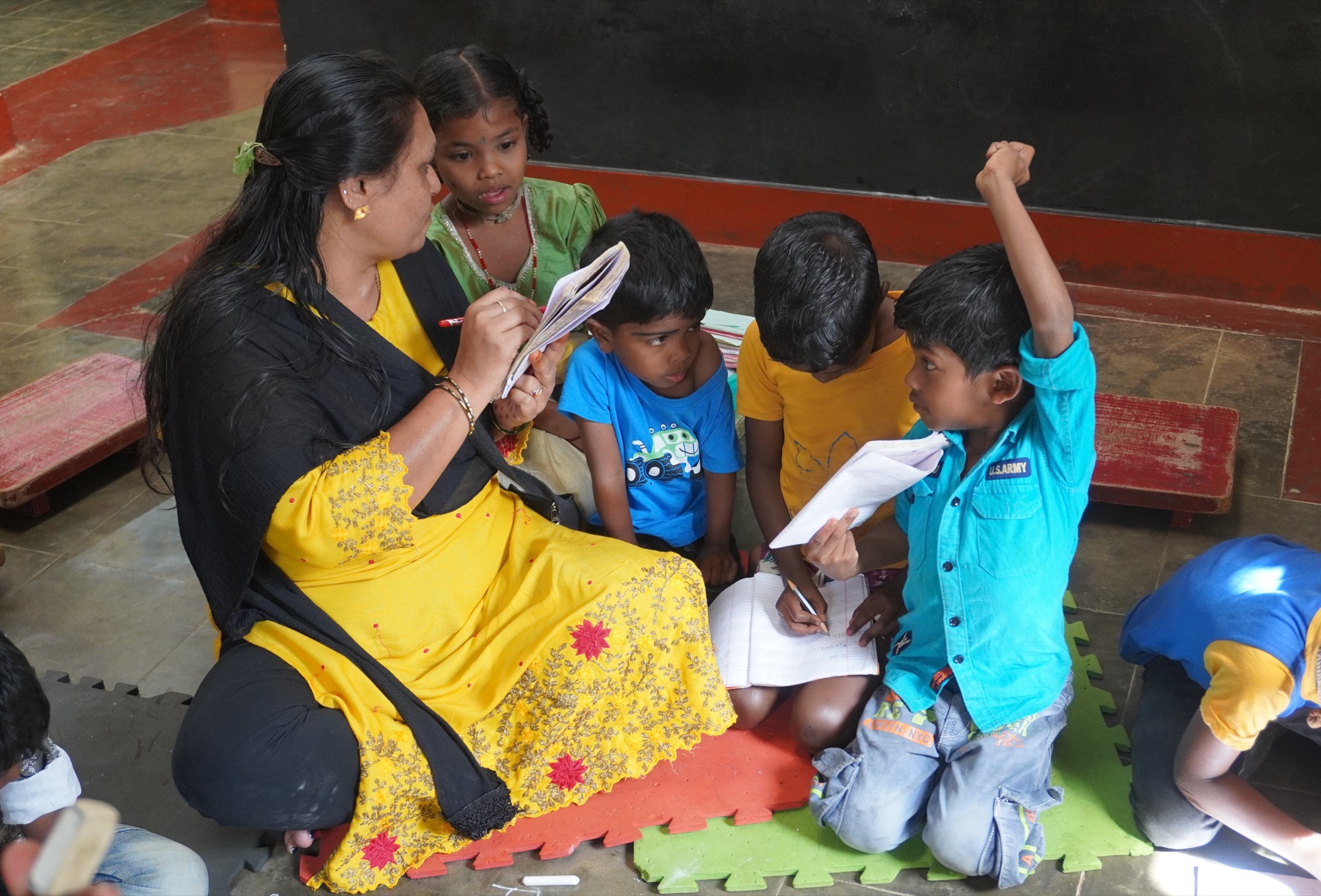 Woman teacher in yellow sari teaching young Adivasi children seated on the floor, a child raising hand eagerly