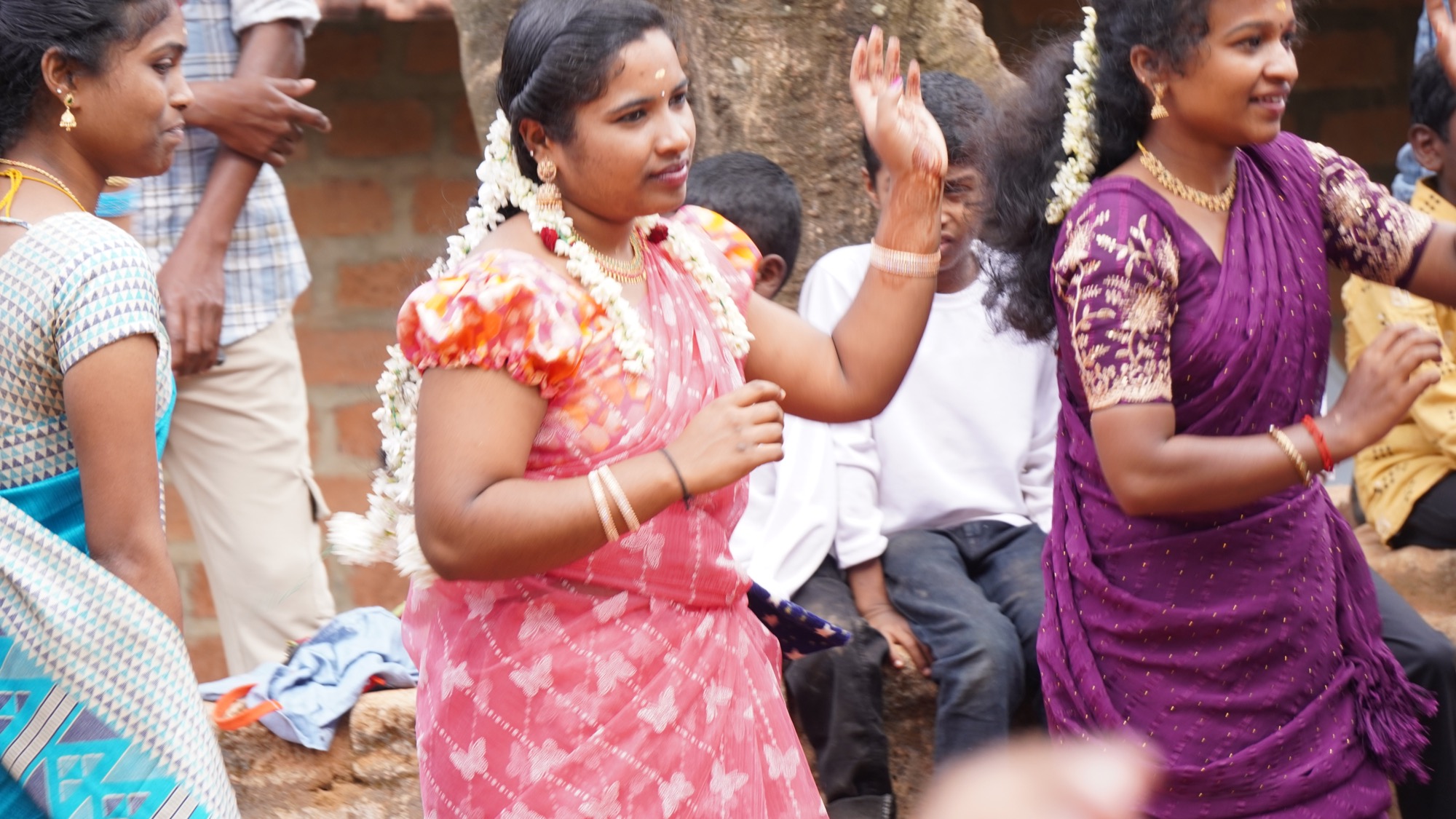 Women in colourful saris dancing energetically in a circle outdoors, children watching