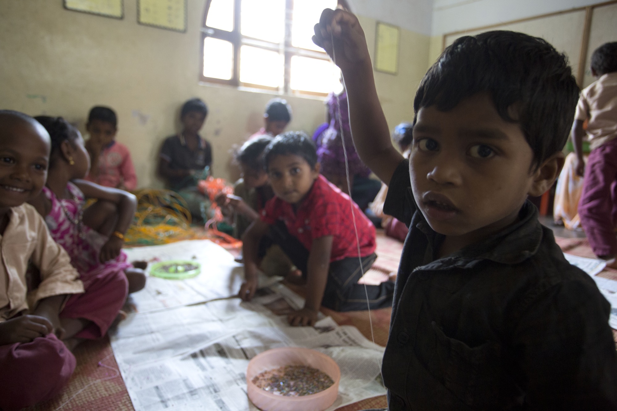 Young Adivasi boy holding up a beading thread during a craft activity, other children working on mats behind